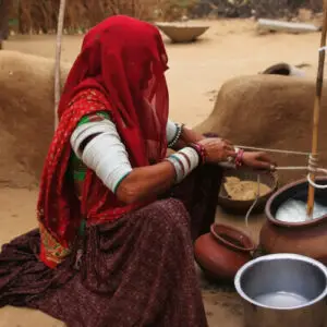A woman in a vibrant red veil and traditional white arm bangles sits on the ground churning milk in a clay pot outside a mud dwelling.