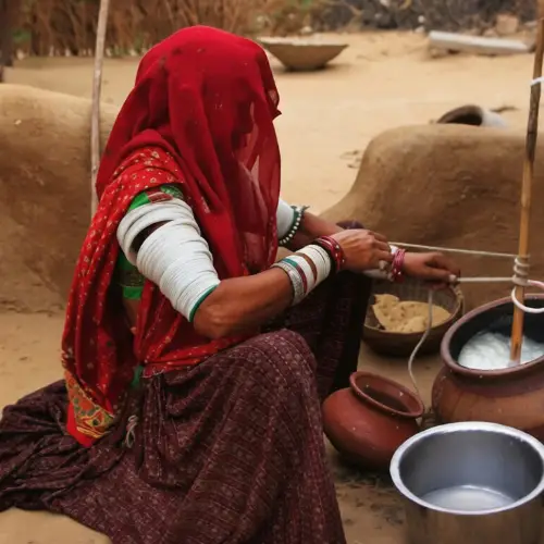 A woman in a vibrant red veil and traditional white arm bangles sits on the ground churning milk in a clay pot outside a mud dwelling.