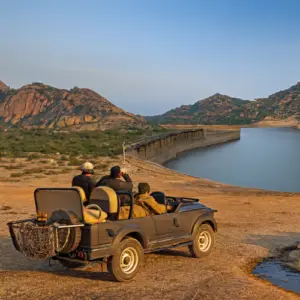 Three people in an open-top safari jeep parked on a rocky overlook, viewing a large curved dam and reservoir nestled between desert mountains under a clear blue sky.