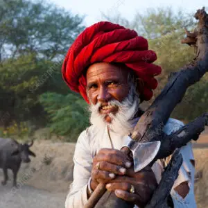 A close-up portrait of an elderly man with a white beard wearing a large, vibrant red turban, holding a wooden staff and a small axe over his shoulder.