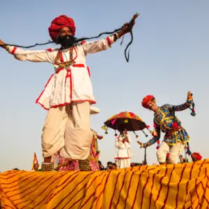 Performers in traditional Rajasthani attire, including colorful turbans and bells, dance on a platform draped in striped yellow cloth under a clear sky.
